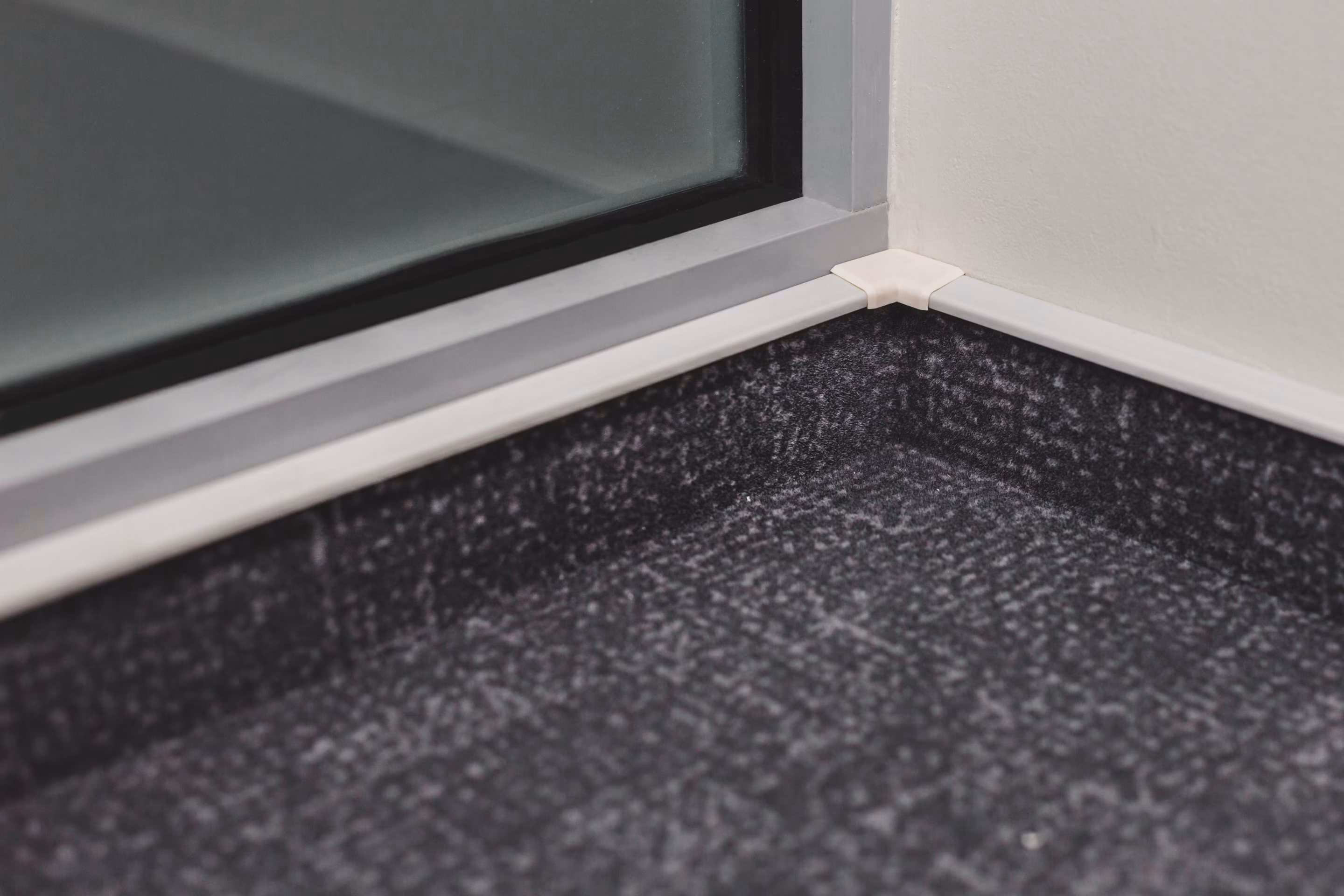 Teppichsockelleiste in einer Innenecke: grauer Teppichboden, an der Wand unter einem weißen Abschlussprofil hochgeführt